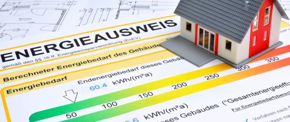 Ein Modellhaus steht auf einem Energieausweis mit Farbskala und detaillierten Energieeffizienzklassen, darüber sind Baupläne sichtbar.
