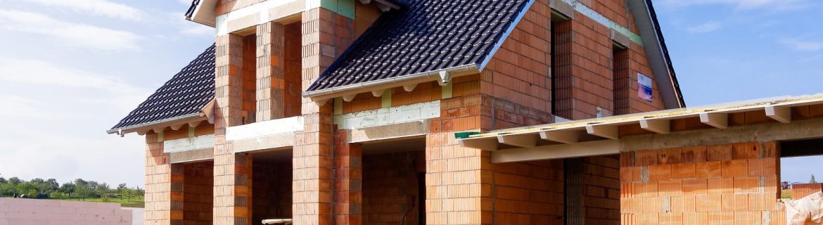 Ein Einfamilienhaus im Rohbau mit Ziegeldach. Die Außenwände sind aus roten Ziegelsteinen errichtet, und die Fenster- und Türöffnungen sind sichtbar.