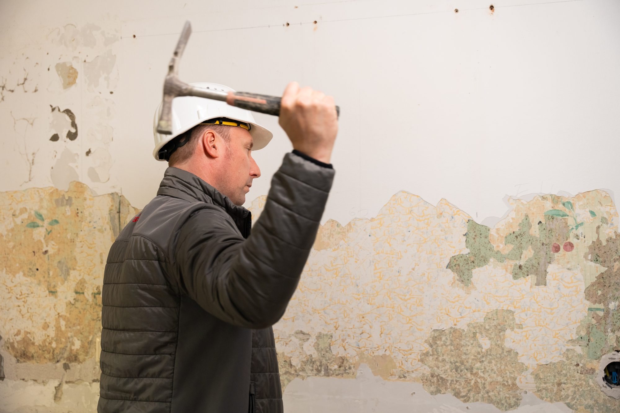 Mann mit Helm und Spitzhammer schlägt gegen eine abgerissene Tapete in einem Raum.