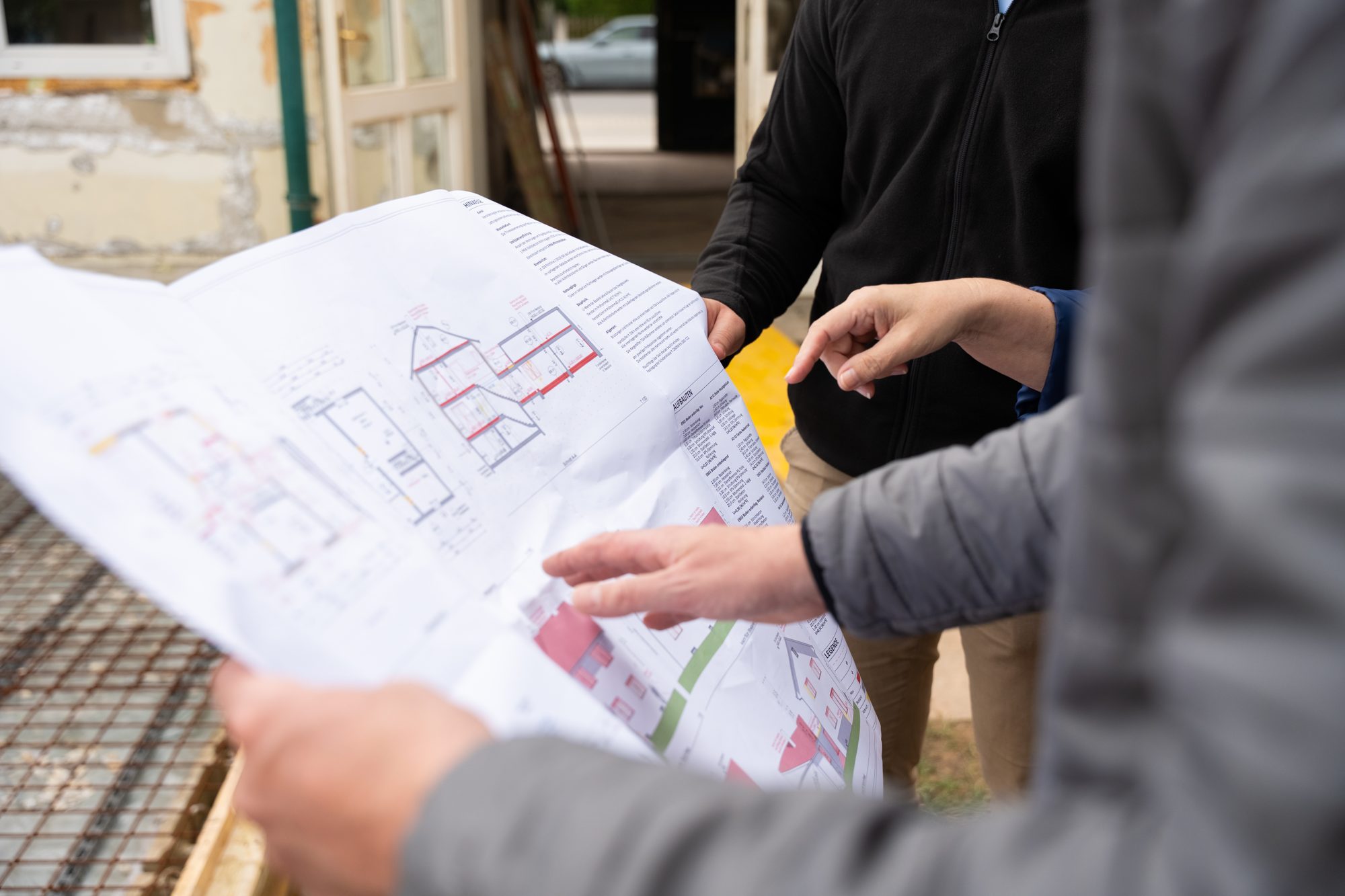 Hände halten einen Bauplan über die verlegte Bewehrung eines Rohbaus, um Plan und Ausführung zu vergleichen