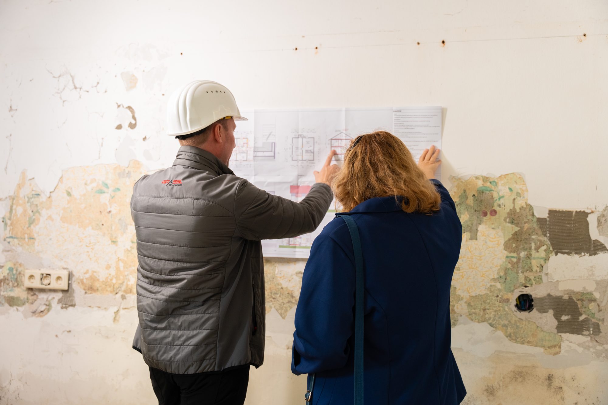 Baumeister Franz Grassl und eine Kundin vergleichen auf der Baustelle einen an die Wand gehefteten Plan mit der Ausführung