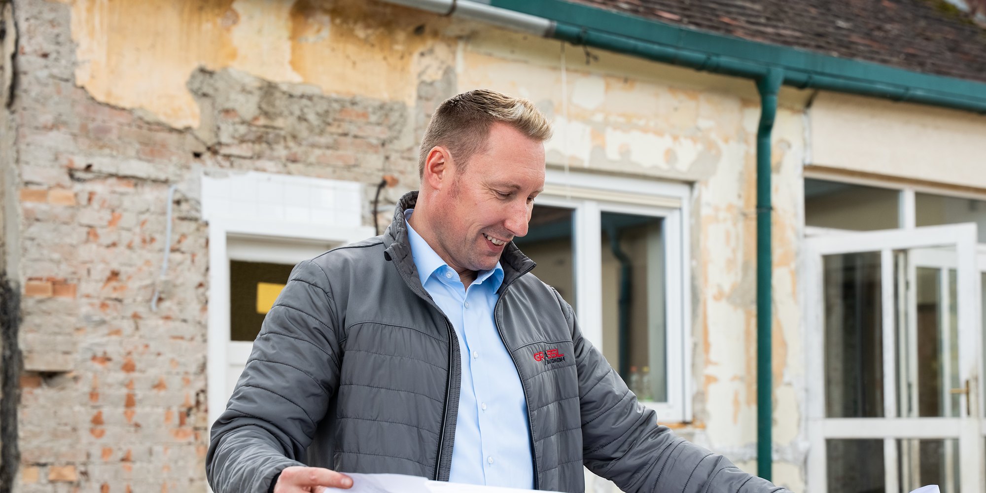 Baumeister Franz Grassl liest auf einer Rohbau-Baustelle vor einem Altbau einen großen Plan, am Boden liegt die Bewehrung